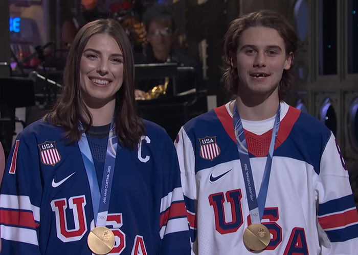Olympic hero Jack Hughes smiling with gold medal alongside teammate in USA hockey jerseys during award ceremony. Olympic hero Jack Hughes smiling with gold medal alongside teammate in USA hockey jerseys during award ceremony.
