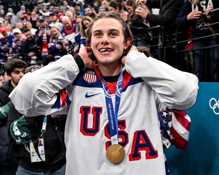 Olympic hero Jack Hughes wearing medal, smiling awkwardly while interacting with an excited crowd after winning gold. Olympic hero Jack Hughes wearing medal, smiling awkwardly while interacting with an excited crowd after winning gold.