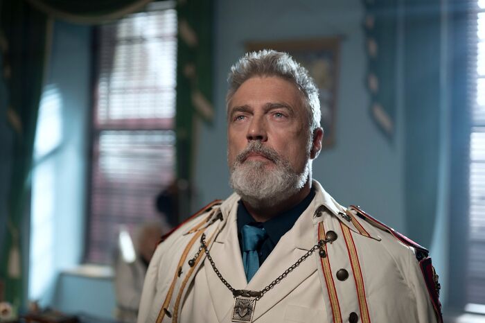 Middle-aged man with gray hair and beard wearing a white uniform in a softly lit room, hinting at a game-changing reveal.