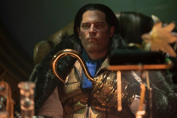 Man dressed in elaborate armor and fur cloak, seated in a dimly lit room, related to One Piece anime and manga continuation.