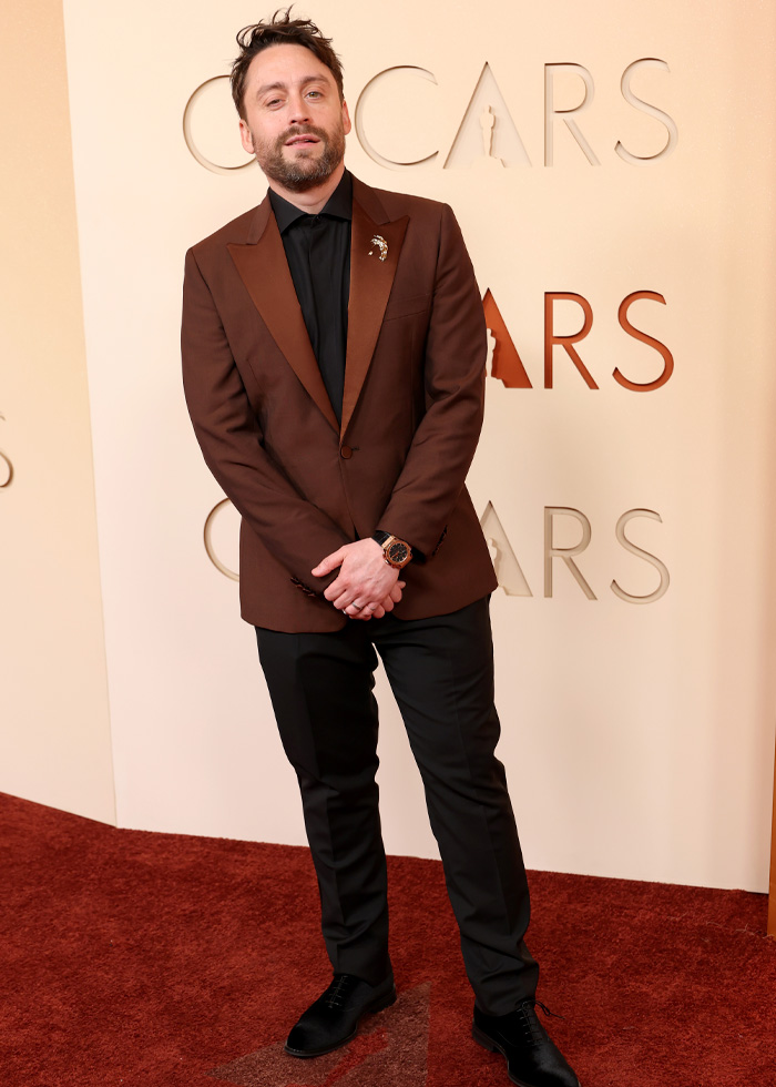 Man wearing a brown tuxedo jacket and black pants, posing on the red carpet showcasing jaw-dropping Oscars 2026 outfits.