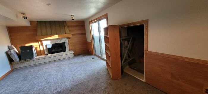 Empty old home living room with fireplace, built-in shelves, and sliding glass door, showing signs of age and neglect.