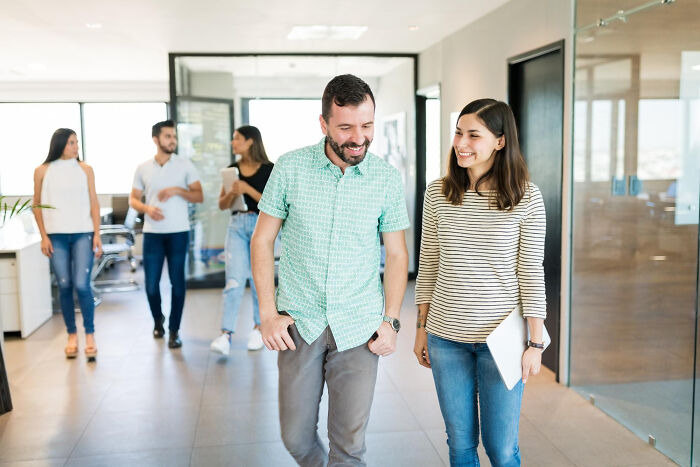 Two colleagues smiling and walking in an office, capturing a savage I literally own the company moment at work.