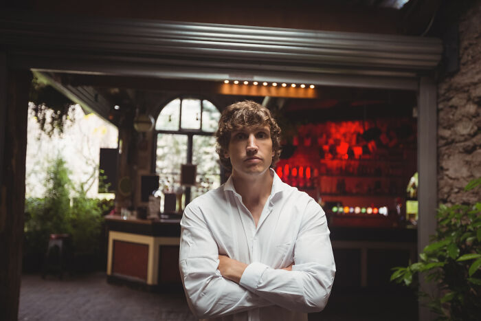 Young man with crossed arms standing confidently inside a dimly lit bar, representing savage I literally own the company moments.