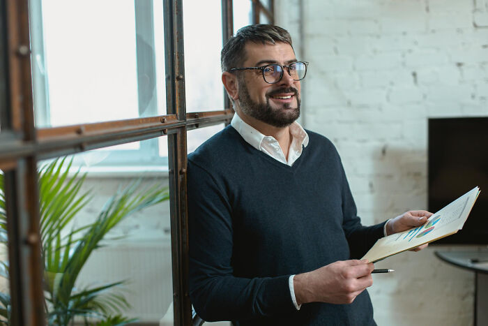 Man with glasses holding documents and pen, smiling confidently during a savage I literally own the company moment.