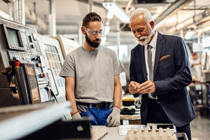 Older businessman and factory worker examining parts together in a manufacturing setting, showcasing company ownership moments.