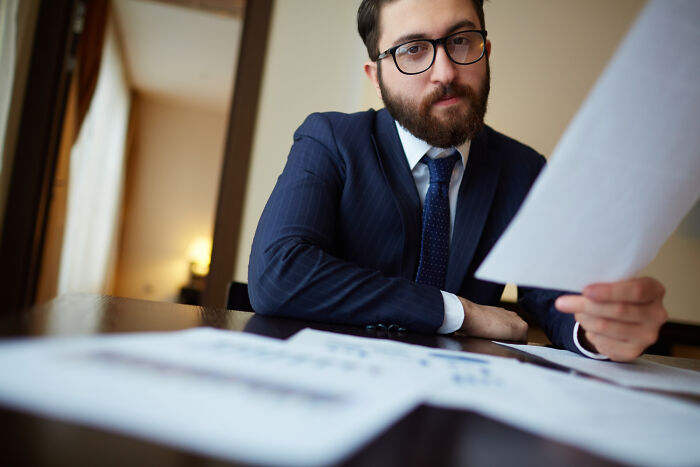 Bearded businessman in glasses reviewing documents at office desk, portraying confident owner company moment.
