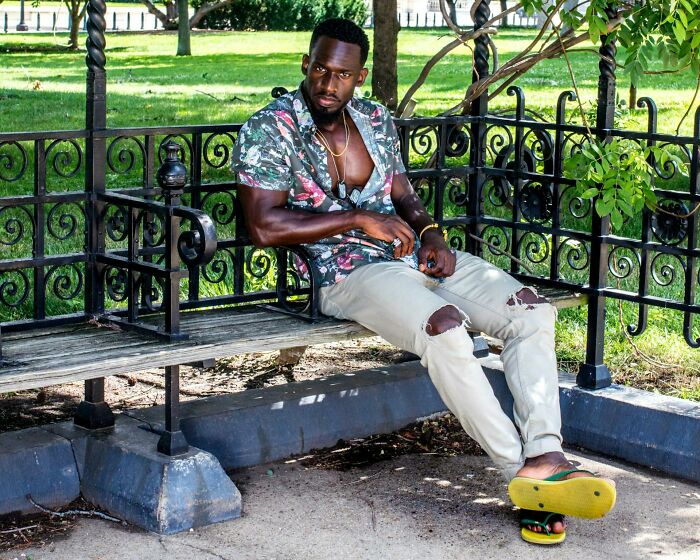 Man in torn pants and floral shirt sitting confidently on a park bench, embodying savage I literally own the company vibes.