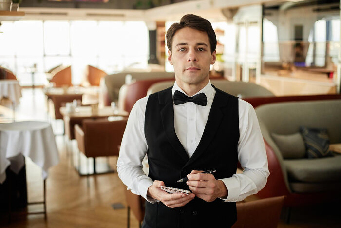 Young waiter in a black vest and bow tie taking an order, representing savage I literally own the company moments