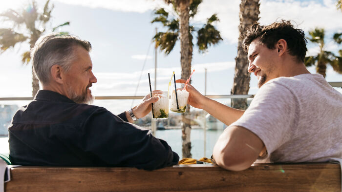 Two men clinking drinks outdoors by the beach, sharing relaxed moments reflecting savage I literally own the company vibes.