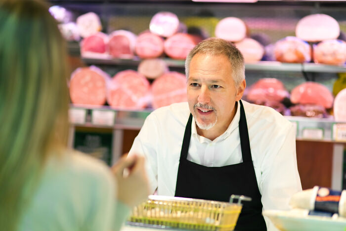 Man wearing black apron serving customer behind deli counter, showcasing savage moments of literally owning the company.