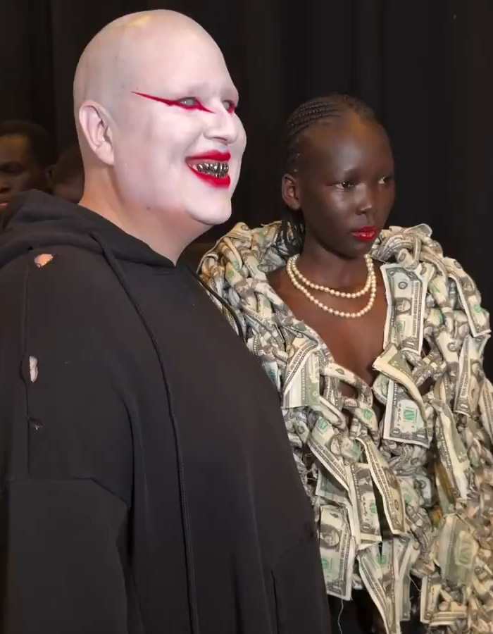 Two models at Paris Fashion Week wearing edgy outfits, one with dollar bill dress and striking makeup inspired by Hunger Games.