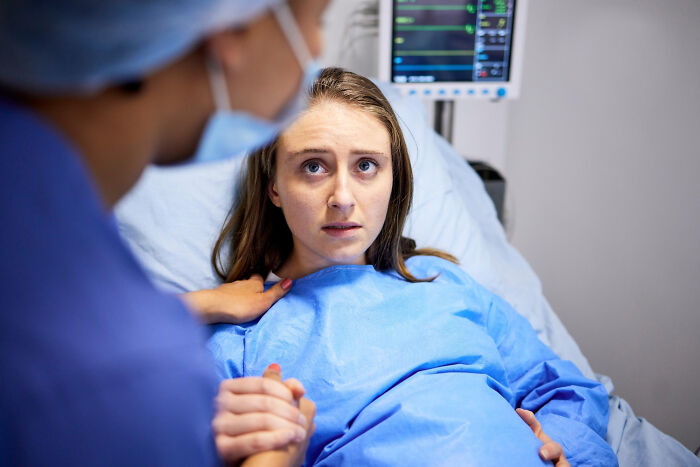 Patient under sedation looks anxious in hospital bed while surgical staff member holds her hand during procedure preparation.