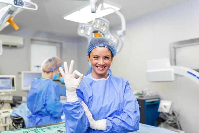 Surgical staff in blue scrubs smiling and making OK sign in operating room during patient sedation procedure.