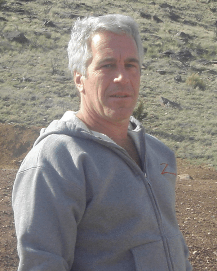 Man wearing a gray hoodie standing outdoors on a rocky and grassy terrain with a serious expression.