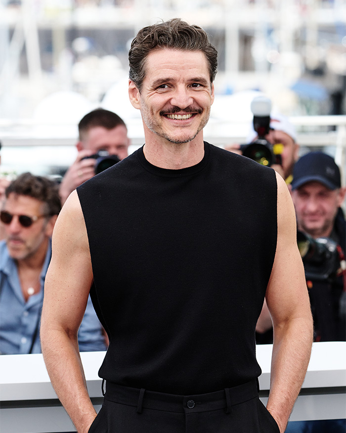 Pedro Pascal smiling in a black sleeveless top at a public event, surrounded by photographers and fans.