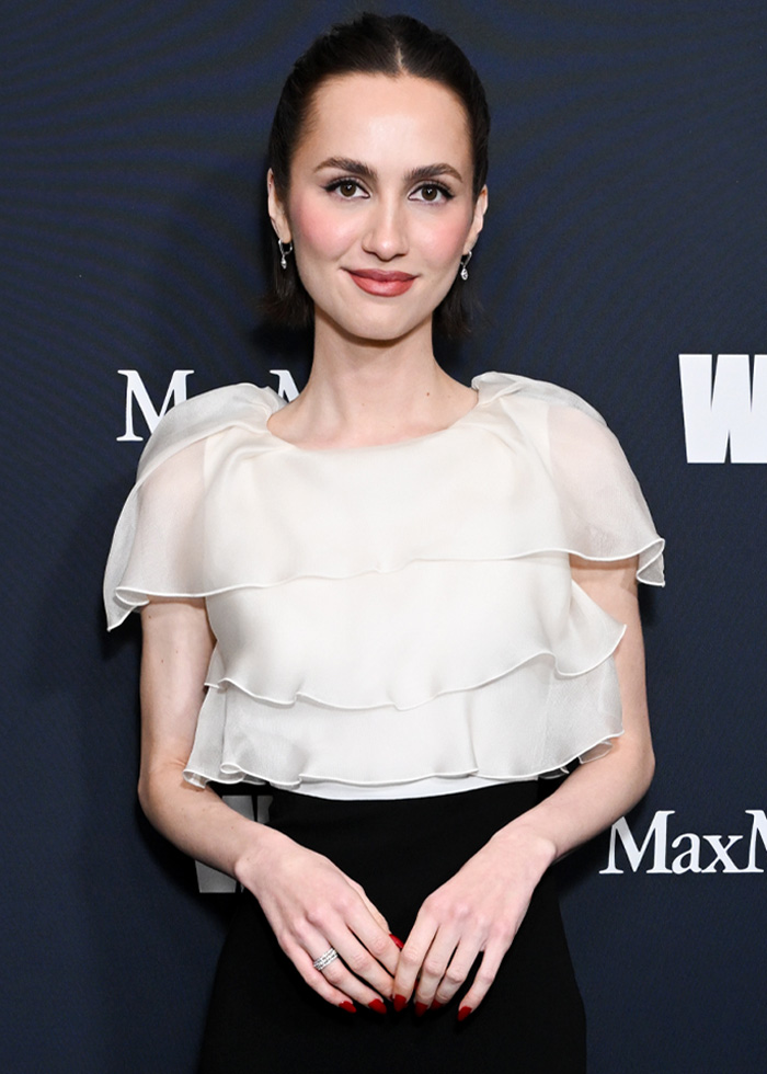 Maude Apatow posing at Oscars party, wearing white ruffled top and black skirt, with a subtle smile.