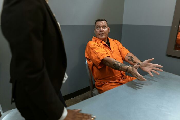 Man in orange prison jumpsuit in a jail interrogation room gesturing with handcuffed hands during questioning about crimes.