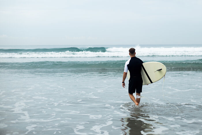 Man holding a surfboard walking into the ocean, reflecting on old petty grudges against strangers he still has.