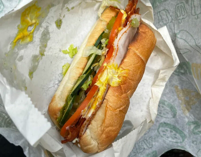 Partially eaten sandwich with bacon, tomato, cucumber, and mustard in a fast food wrapper on a table surface.