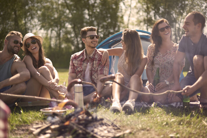 Group of friends sitting by a campfire outdoors, sharing laughs and enjoying a casual social gathering in nature.