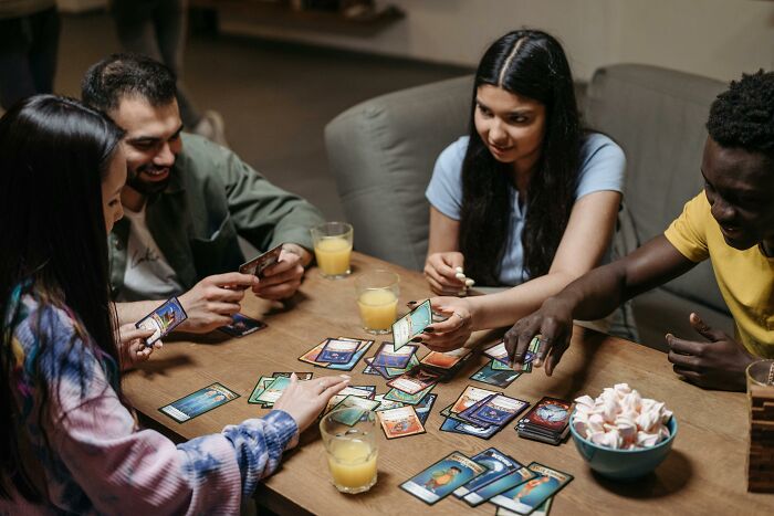 Group of friends playing a card game at a table, showing expressions of focus and enjoyment with drinks nearby.