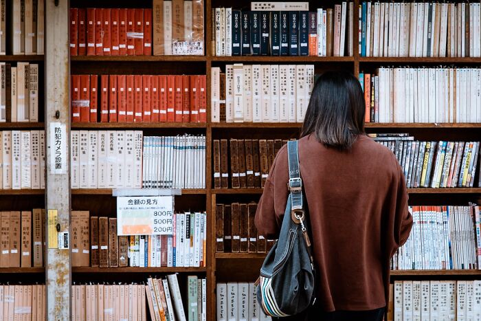 Woman with shoulder bag browsing books on wooden shelves, surrounded by a collection of bizarre true stories and unusual books.