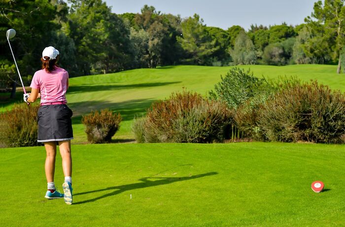 Woman golfer in pink shirt swinging club on a green golf course with trees and bushes in the background, bizarre true stories concept.