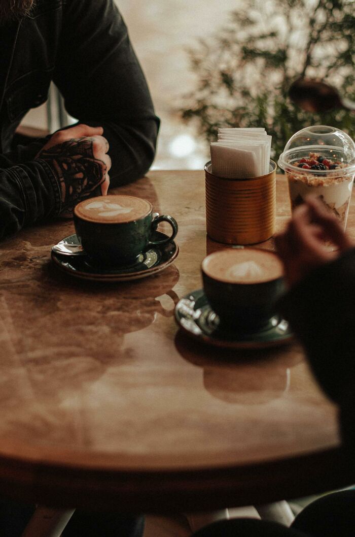 Two people with tattoos having coffee at a table, capturing a moment of serious conversation that sounded like a joke.
