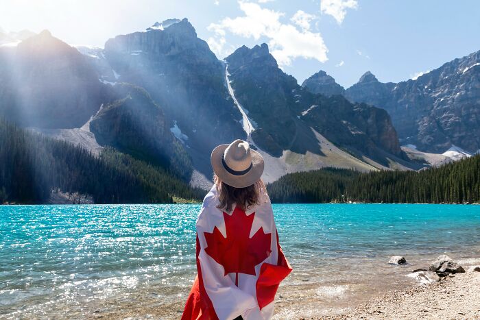 Person wrapped in a Canadian flag by a turquoise lake with mountains, representing vanished truths from home countries.