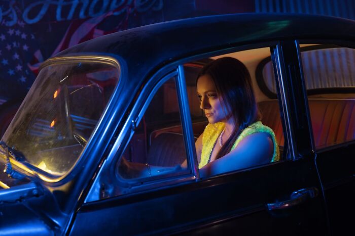 Young woman sitting in a vintage car at night, illuminated by blue and orange lights, capturing a reflective moment.