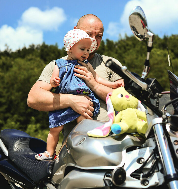 A dad holds his toddler daughter on a motorcycle seat, showing wholesome support and care in an outdoor setting.