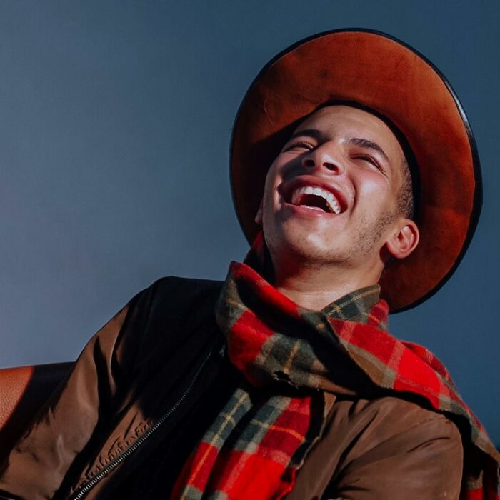 Young man laughing loudly wearing a brown cowboy hat and plaid scarf, capturing the vibe of disastrous first dates.