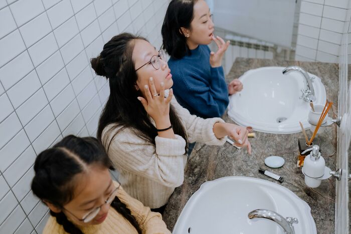 Three women in a bathroom applying makeup and skincare, illustrating secrets women don't tell men in personal care routines.