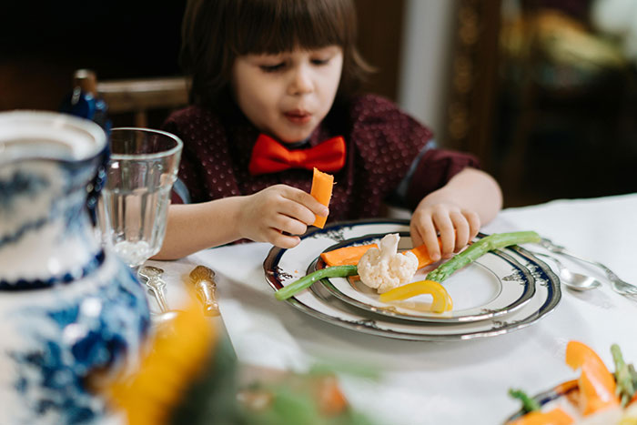 Young child with a red bow tie eating fresh vegetables at a dining table, highlighting vegan-friendly meal choices.