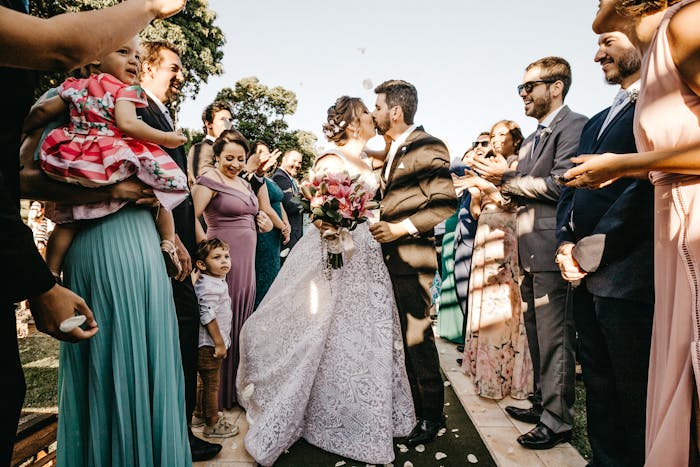 Couple kissing at sister&rsquo;s wedding aisle while guests in formal outfits watch and celebrate outdoors in daylight.