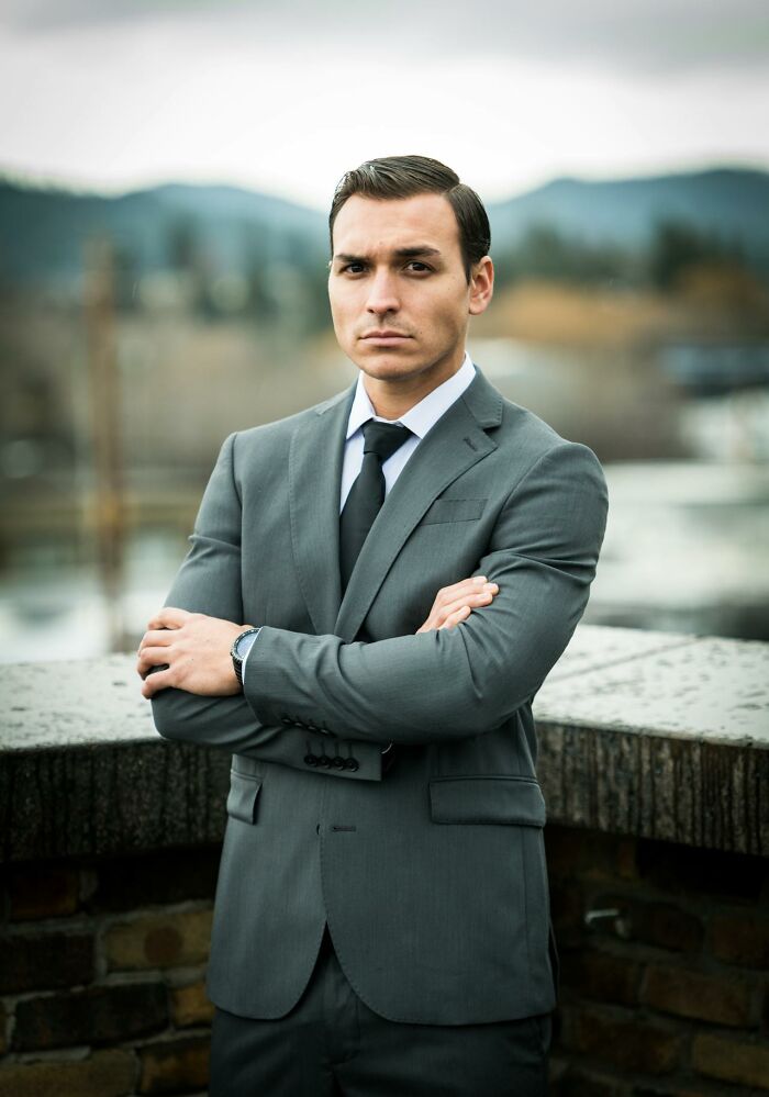 Serious man in a gray suit with arms crossed, outdoors with blurred landscape, reflecting things people said sounding like a joke.