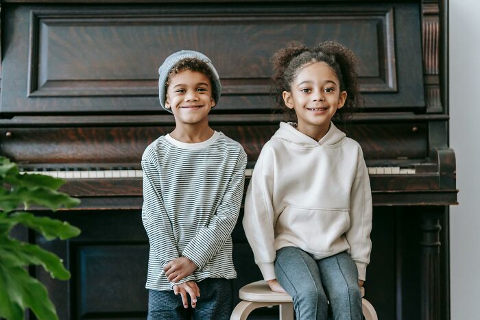 Two smiling children in casual clothes posing in front of an old piano for a celebrity childhood quiz image.