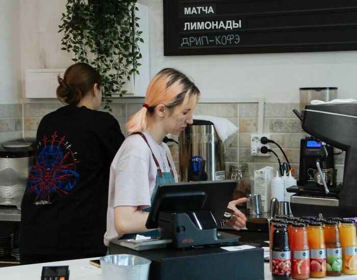 Two cafe workers preparing drinks behind the counter with a menu board and juice bottles, capturing Gen Z stare reactions.