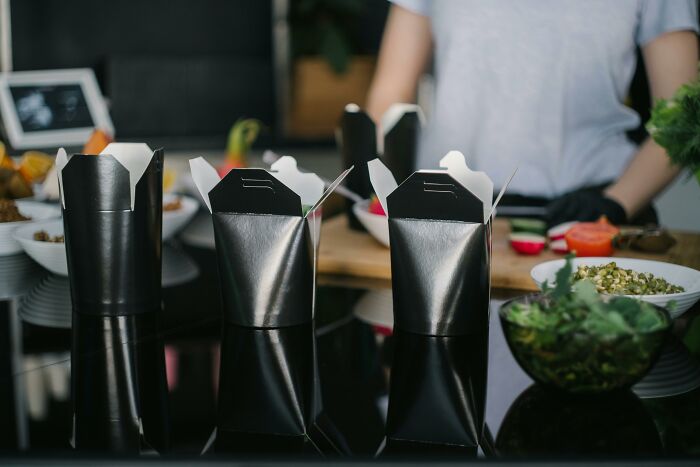 Three black takeout containers on a reflective surface with a person preparing food, illustrating ridiculous customer complaints.