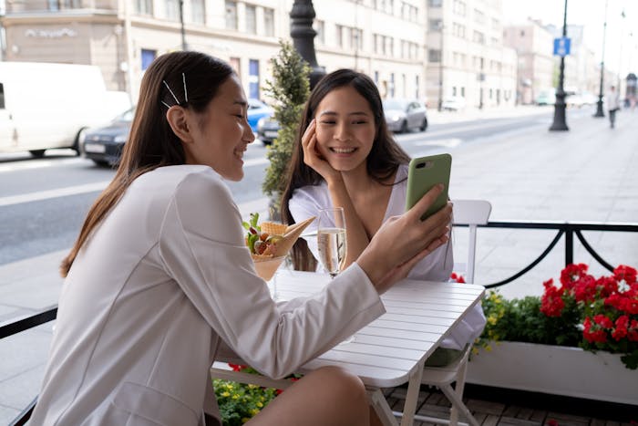 Two women sitting at an outdoor cafe, smiling and looking at a phone, discussing outfit choices for sister&rsquo;s wedding drama.