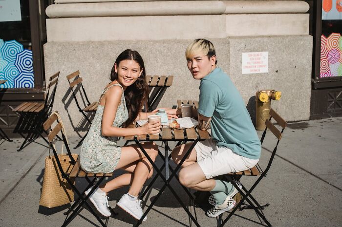 Young couple sitting at outdoor cafe table on a casual first date, enjoying coffee and conversation under sunny weather.