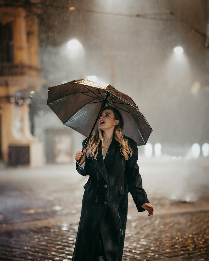 Young woman holding umbrella in rainy street at night, capturing moments that sound like a complete joke but are serious.