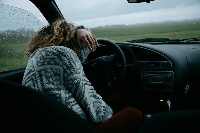 Person sitting in a car with head resting on the steering wheel, reflecting on forgiving a cheating spouse during a rainy day.