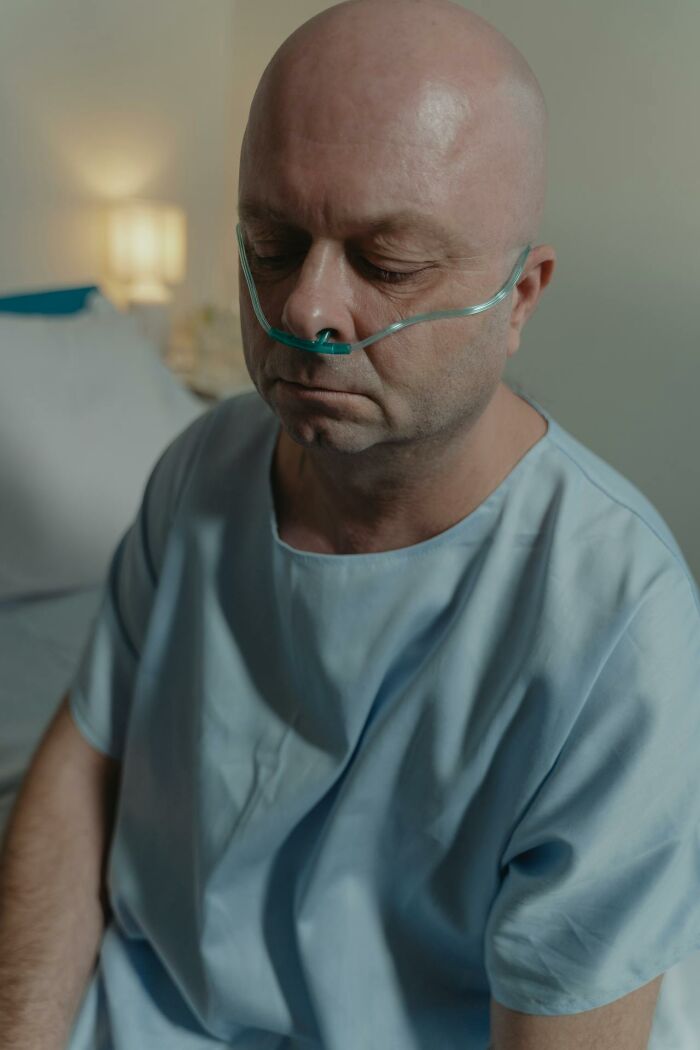 Middle-aged man in hospital gown with nasal oxygen in a dimly lit room, reflecting on ER medical staff experiences.