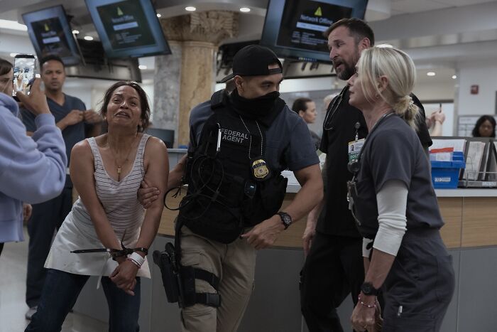 A distressed woman being held by a federal agent in a busy office setting for The Pitt season 2 guest stars.