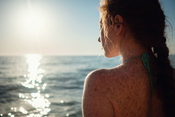 Young woman with freckled skin gazes at ocean sunset, illustrating unusual aspects that freak people out about their bodies.