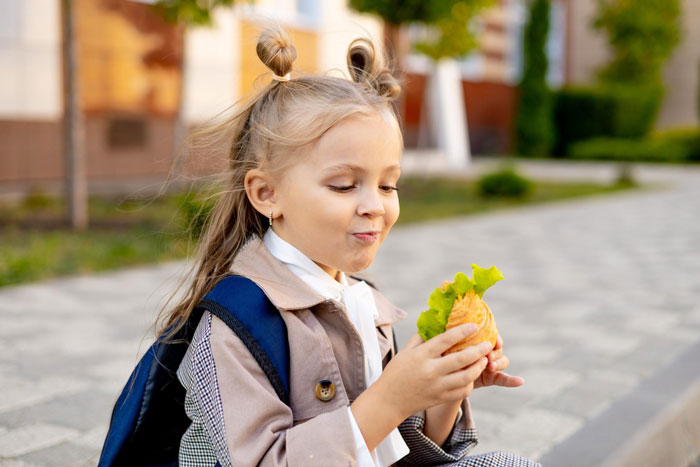 Young schoolgirl enjoying a sandwich outdoors with a backpack, illustrating the refuse to stop making school lunches concept.