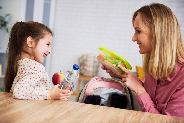 Mother packing a healthy school lunch while smiling at her daughter, highlighting the importance to refuse stop making school lunches.