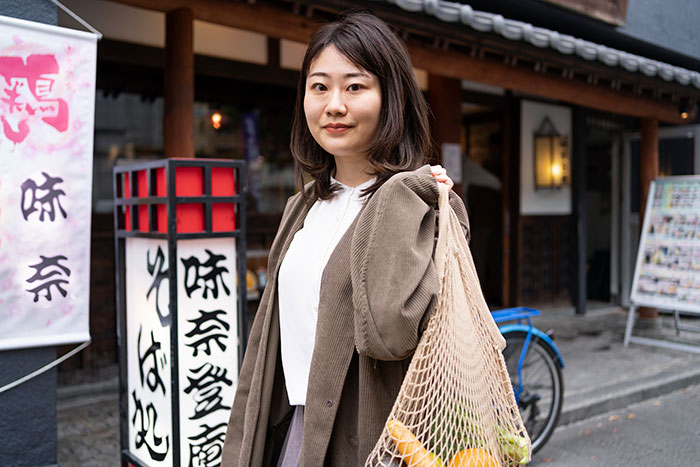 Young woman standing outside a traditional building, carrying a net bag, embodying the theme of popped the question embarrassment.